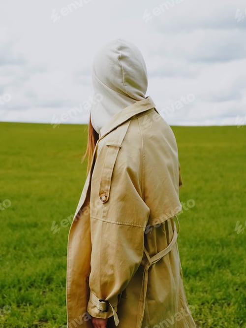Preview: Silhouette of a girl in a hood in beige colors against the background of green grass and sky