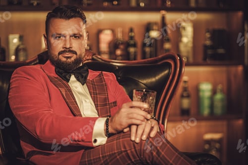 Preview: Extravagant stylish man with whisky glass sitting on armchair in