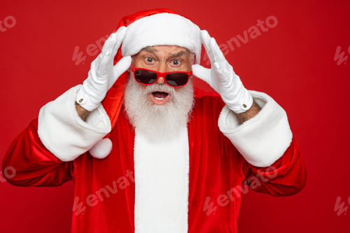 Preview: Amazed shocked surprised Santa Claus face in sunglasses with open mouth isolated on red background