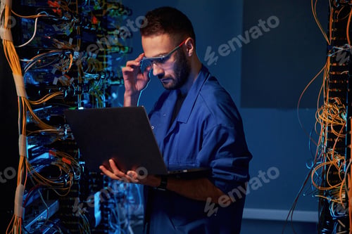 Preview: Blue colored lighting. Young man is working with internet equipment and wires in server room
