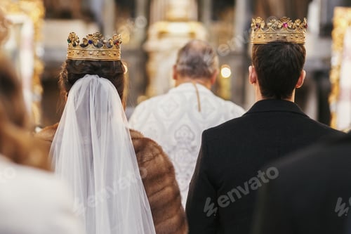 Preview: gorgeous bride and stylish groom in golden crowns, standing with priest in church