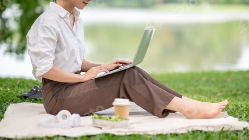 Preview: Close up of a woman in white shirt using laptop while sitting knee up on picnic mat laying on grass.