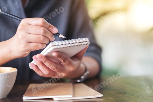 Preview: Closeup image of a woman holding and writing on blank notebook with coffee cup on table in cafe