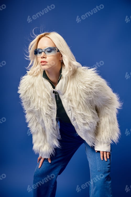 Preview: beautiful woman in faux fur jacket, jeans and sunglasses posing on blue backdrop, winter fashion