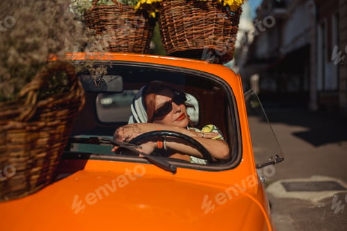 Preview: Young beautiful woman in a silk scarf and sunglasses sits behind the wheel of an orange retro car