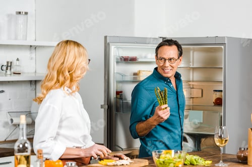 Preview: mature wife cutting vegetables and husband holding asparagus in kitchen