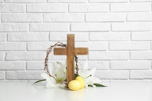 Preview: Wooden cross, crown of thorns, Easter eggs and blossom lilies on table against brick wall