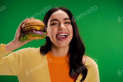 Preview: brunette woman holding tasty burger and sticking out tongue isolated on green