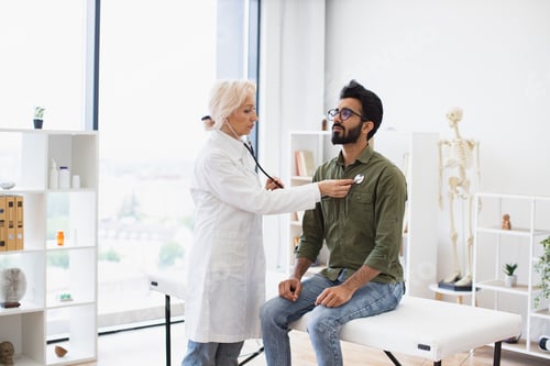 Preview: Elderly lady using stethoscope while young man sitting on exam couch.