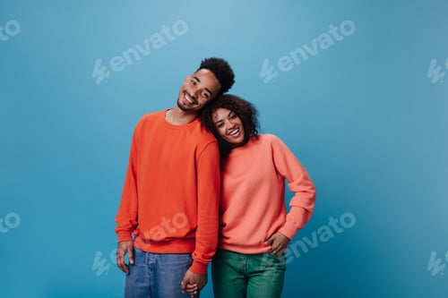 Preview: Couple in love looking into camera on blue background. Portrait of happy curly woman in orange swea