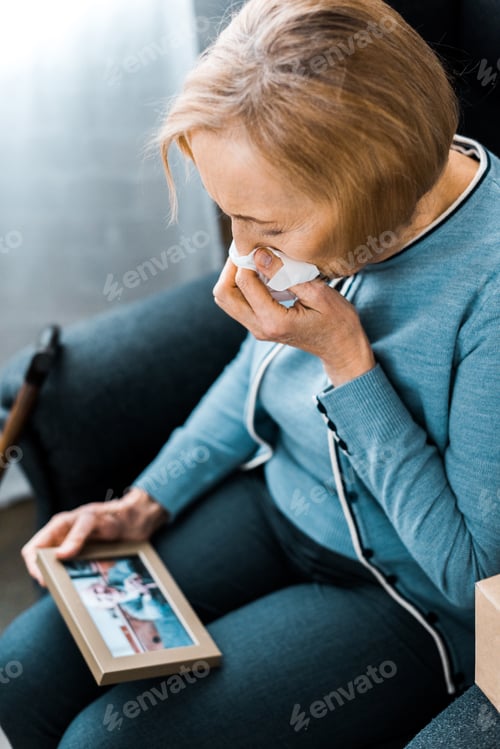 Preview: sad senior woman sitting and crying while looking at picture frame