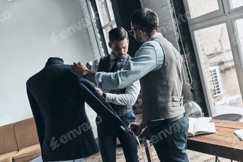 Preview: Best designs. Two young fashionable men measuring jacket?s sleeve while standing in workshop