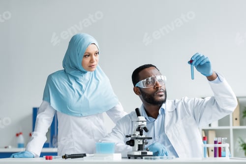 Preview: Interracial scientists in latex gloves looking at sample near microscope in laboratory