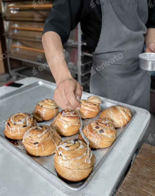 Preview: A young Hispanic baker is pouring sugar glaze on some baked cinnamon rolls