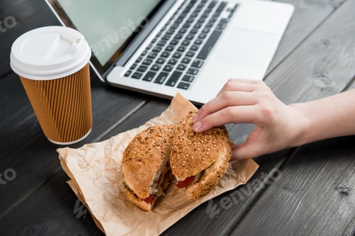 Preview: close up of hand holding burger with coffee to go and laptop on wooden tabletop