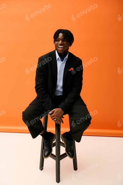 Preview: Handsome young man smiles joyfully while sitting on a stool against a vibrant orange background