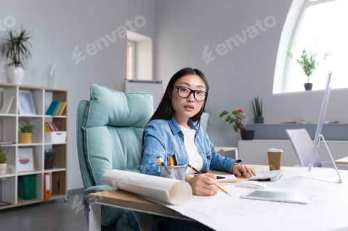 Preview: Portrait of a young Asian woman architect in glasses working at a computer. Looks into the camera