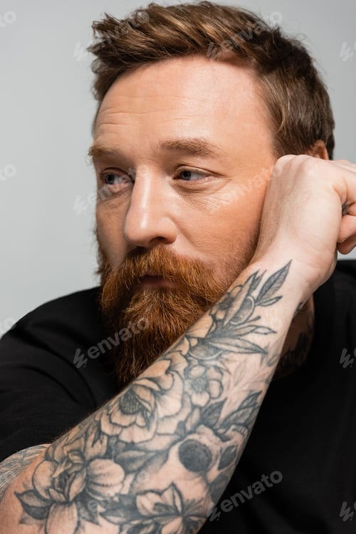 Preview: portrait of thoughtful bearded man holding tattooed hand near face and looking away isolated on grey