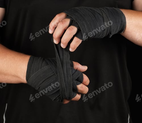 Preview: Adult athlete stand in black clothes and wrap his hands in black textile elastic bandage