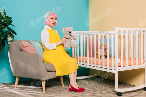 Preview: retro styled pregnant pin up woman with pink hair sitting with teddy bear near baby cot in child