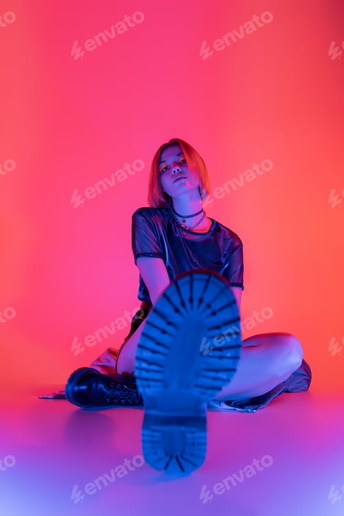 Preview: young woman in trendy boots looking at camera while sitting on purple and coral background