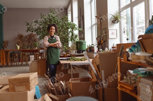 Preview: Attractive smiling florist stands with crossed arms on background of her own floral decor studio
