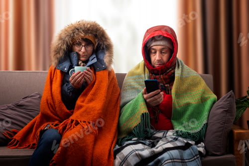 Preview: Middle age family in parkas drinking coffee on the sofa while using smartphone in their cold living
