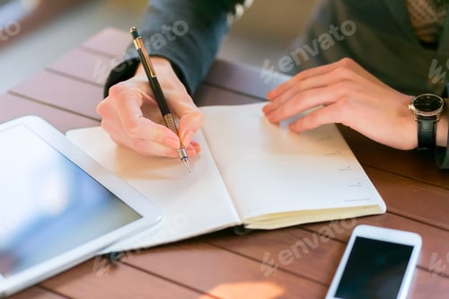 Preview: Person writing in a notebook at a wooden table in a bright workspace with digital devices nearby