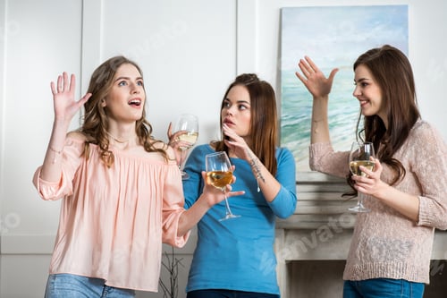 Preview: Three excited young attractive women holding glasses with white wine and gesticulating