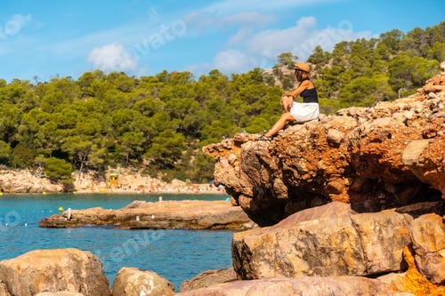 Preview: Vacation concept, A young woman at Salada y Saladeta beach sitting on some rocks by sea. Balearic