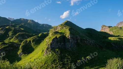 Preview: Lush green mountains under a clear blue sky with scattered clouds on a sunny day in Riale, Italy