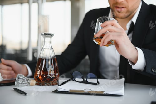 Preview: Fatigue young businessman sitting at workplace and holding whiskey bottle, drinking alcohol