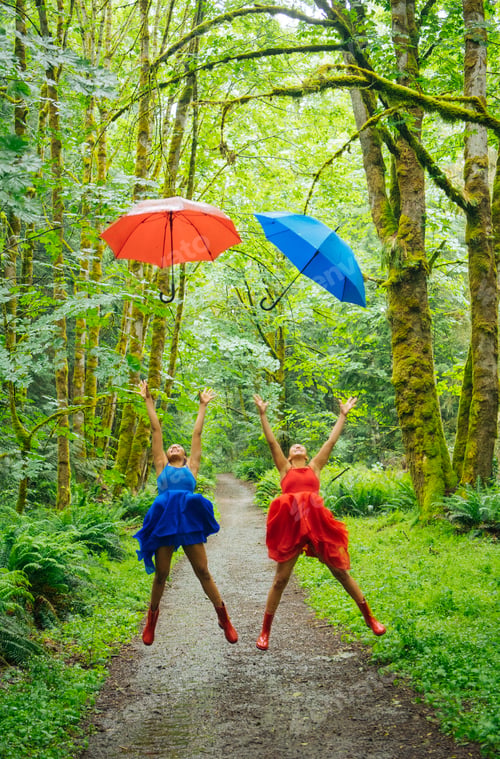 Preview: Two joyful individuals jumping with colorful umbrellas on a forest path surrounded by green foliage.