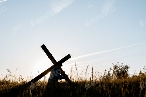 Preview: Male carrying a hand made wooden cross in a grassy field with a blue sky in the background