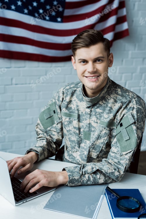 Preview: cheerful soldier looking at camera while using laptop in office