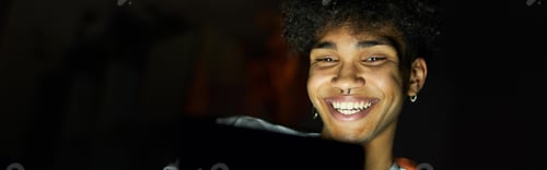 Preview: Portrait of joyful young guy smiling while using tablet pc at night, sitting in dark