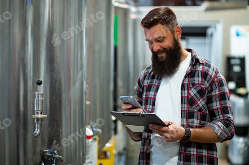 Preview: Man working in brewery and taking notes of the beer production and using smartphone