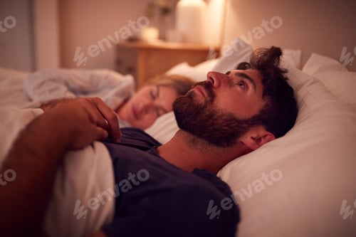 Preview: Couple With Man Lying In Bed Awake At Night Suffering With Insomnia