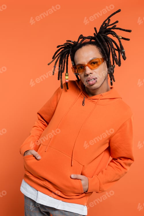 Preview: pierced multiracial man in hoodie and stylish sunglasses posing with hands in pockets on coral
