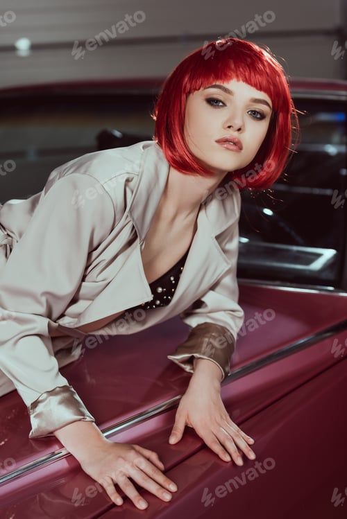 Preview: beautiful young woman in red wig and stylish trench coat looking at camera while leaning at classic