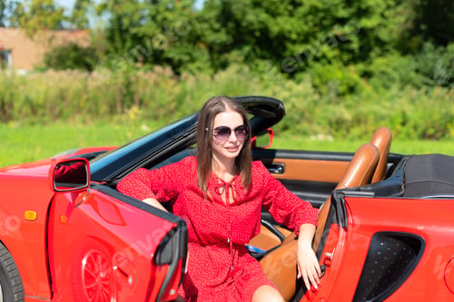Preview: Lifestyle portrait of a beautiful woman in red dress and sunglasses sitting on red cabriolet car
