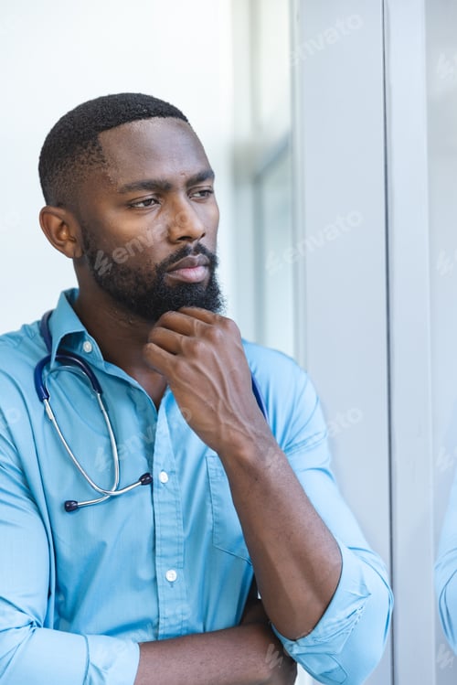 Preview: Thoughtful african american male doctor wearing blue shirt looking through window in hospital room
