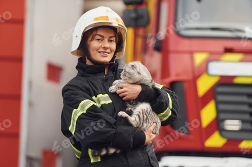 Preview: Standing against big truck and holding beautiful cat. Woman firefighter in uniform is at work