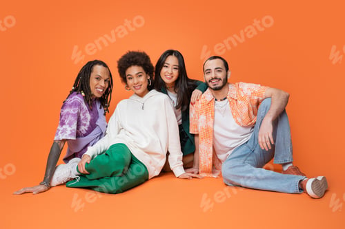 Preview: joyful multicultural friends in stylish clothes sitting and looking at camera on orange background