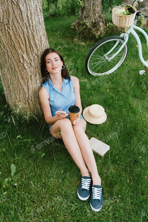 Preview: high angle view of beautiful woman smiling while holding smartphone and paper cup near tree trunk