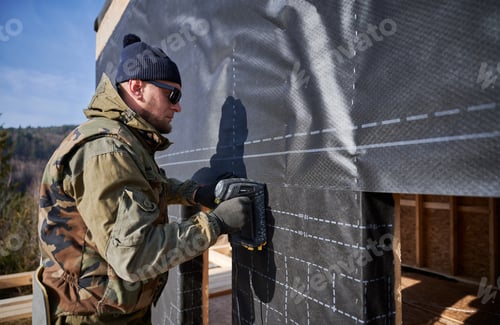 Preview: Male builder installing waterproof membrane on wall of future wooden frame house.