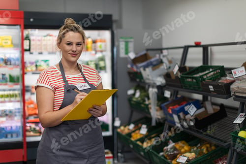 Preview: A young shop assistant in supermarket standing and writing in front of vegetable section.