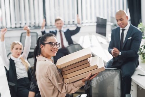 Preview: young businesswoman holding pizza boxes and smiling at camera while coworkers sitting behind in open