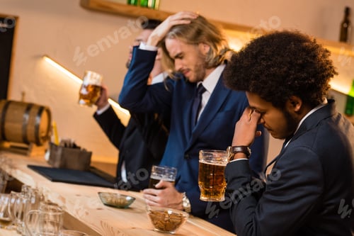 Preview: upset young multiethnic businessmen drinking beer together in bar after work