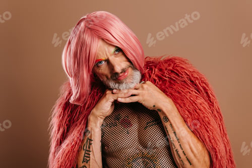 Preview: Bearded mature gay man in pink wig leaning face on hands against brown background